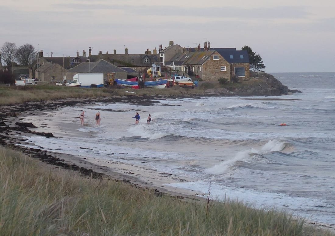 Cuatro bañistas metiéndose en el mar entre olas en una playa de invierno, con cielo gris y el pueblo al fondo