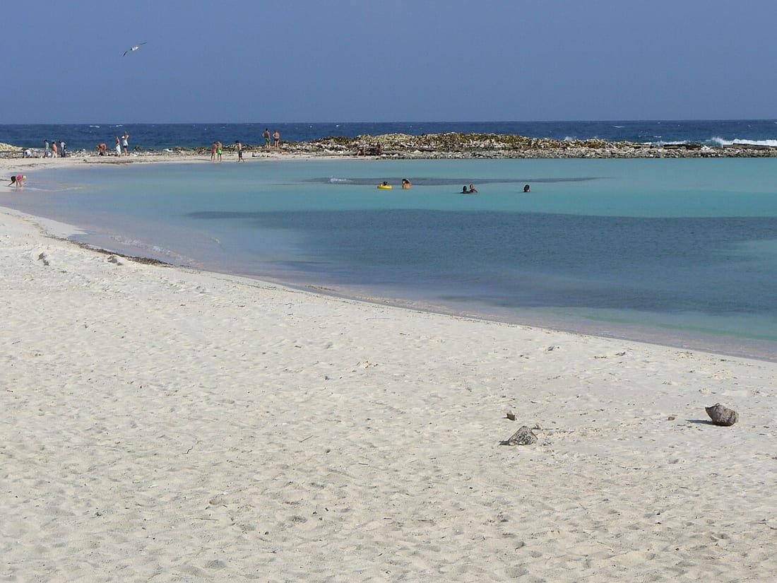 Playa de arena blanca y laguna turquesa poco profunda con bañistas, ideal para un bebé en la playa