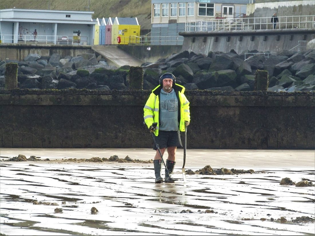 Persona rastreando con un detector de metales la arena mojada de una playa en bajamar, con auriculares y una pala de mano