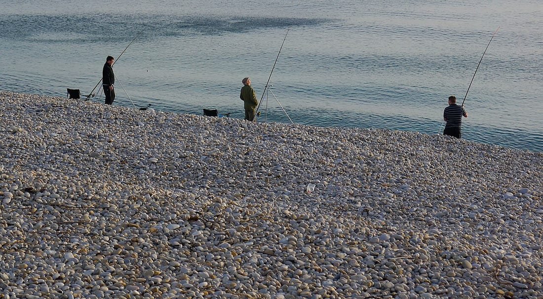 Tres pescadores con cañas largas de surfcasting plantadas en la orilla de una playa de cantos rodados