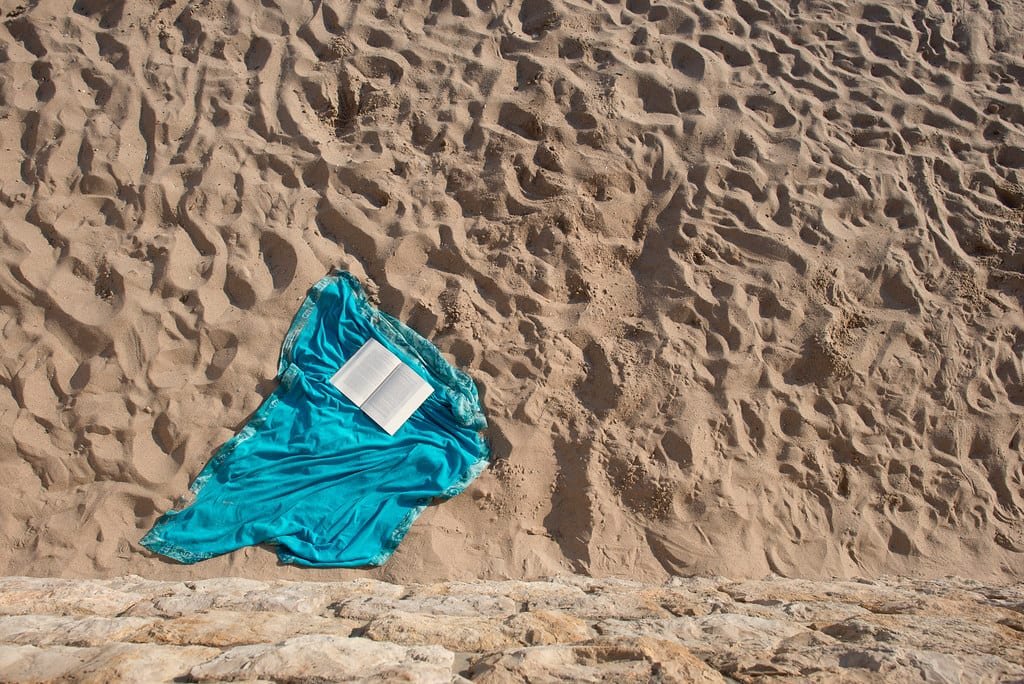 Libro abierto sobre un pareo turquesa extendido en la arena de la playa, visto desde arriba